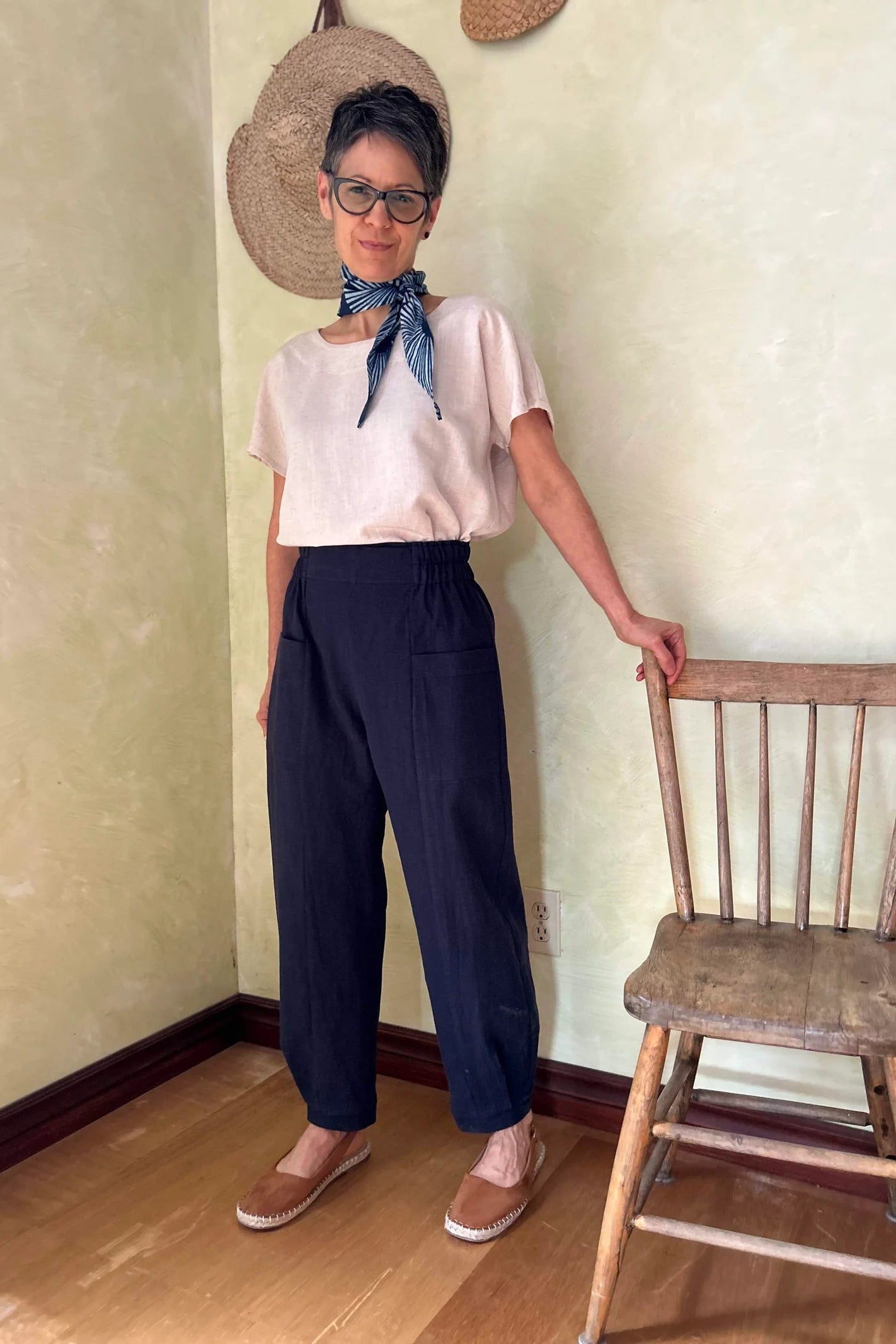 A woman wearing the Oxalis Pants from Kazak in Indigo, with a flat front waistband, legs that taper at the ankle, and large patch pockets. She is wearing them with a cream top and standing in a room with a wooden chair. 