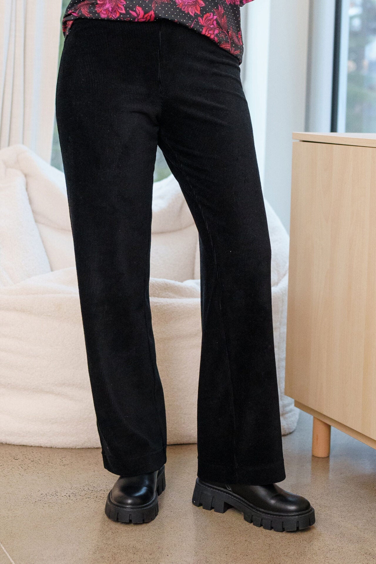 A woman wearing the Carex Pants by Rien ne se Perd in Black, relaxed fit pants with an elastic waist in stretchy corduroy. She is wearing them with a printed top and standing indoors. 