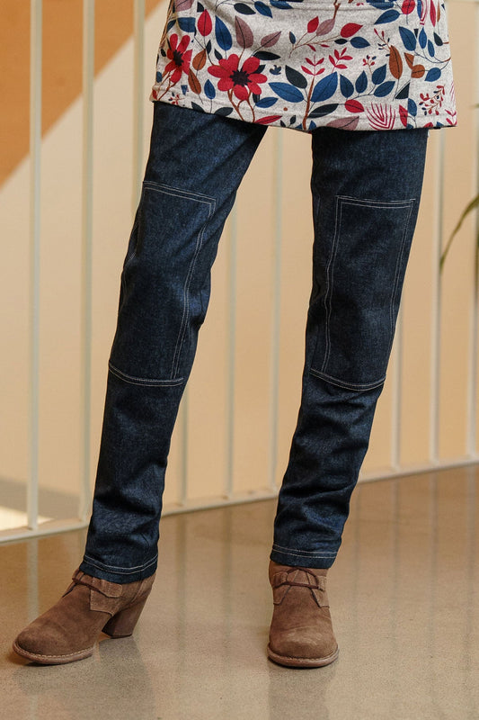 Waist down view of a woman wearing the Cypres Pants by Rien ne se Perd in Blue Denim, stretchy faux denim pants with a pull on waist and tone-on-tone patches on the knees. She is wearing with Genevrier Tunic and standing in front of a railing. 