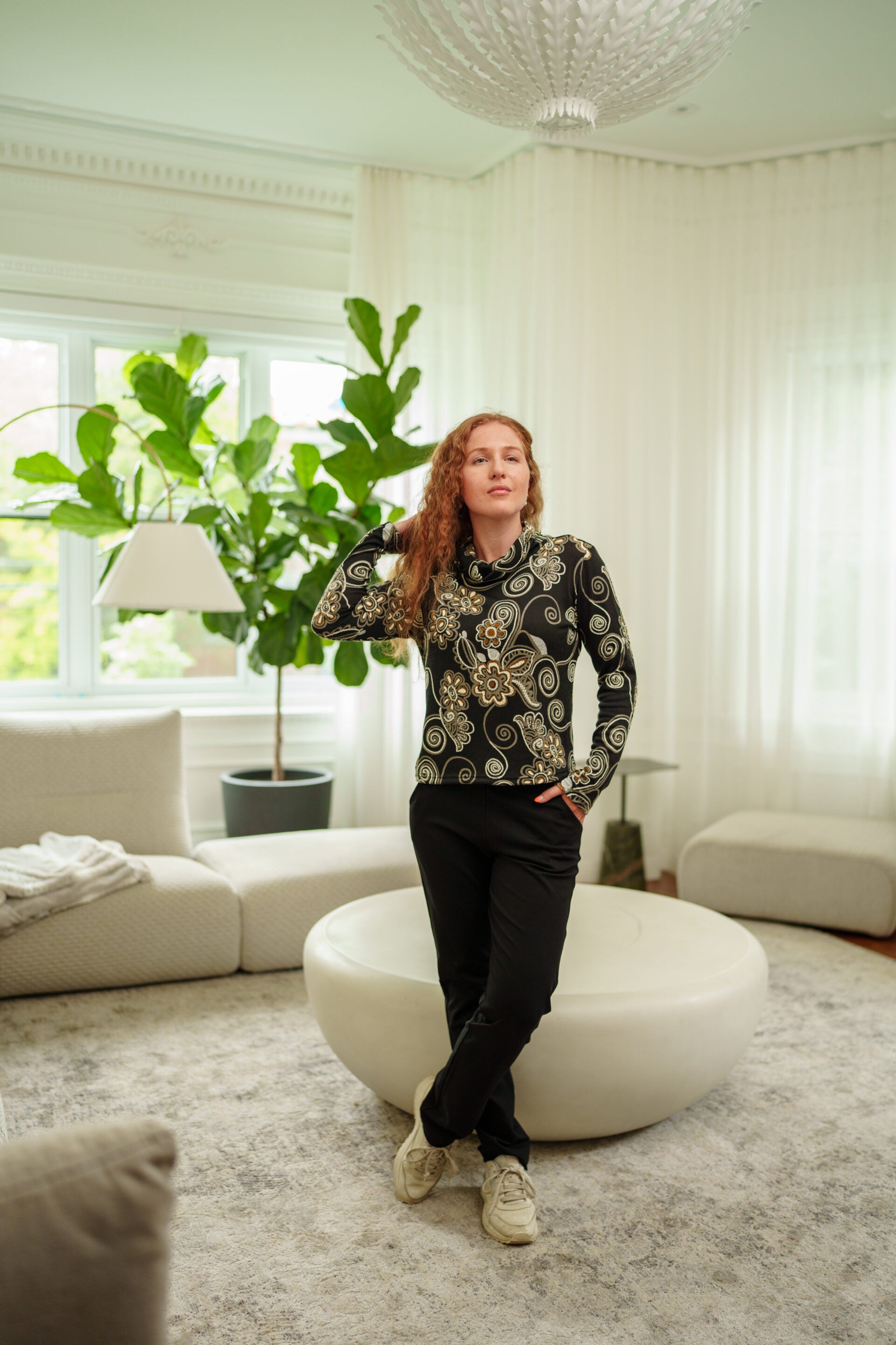 A woman wearing the Murphy Pants by  Rien ne se Perd, pull-on pants with a smooth front waistband, elastic at the back, and a slim fit with a bootcut hem. She is wearing them with black long-sleeve top with gold and white swirl patterns, and beige sneakers, standing in a modern living room with a round white ottoman.