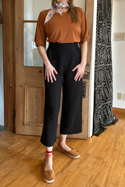 A woman wearing the Petunia Pants by Kazak in Black, wide legged cropped pants with a high waist that's smooth in front with elastic at the back, and large patch pockets. She is wearing them with an orange top and standing in front of a wooden door. 