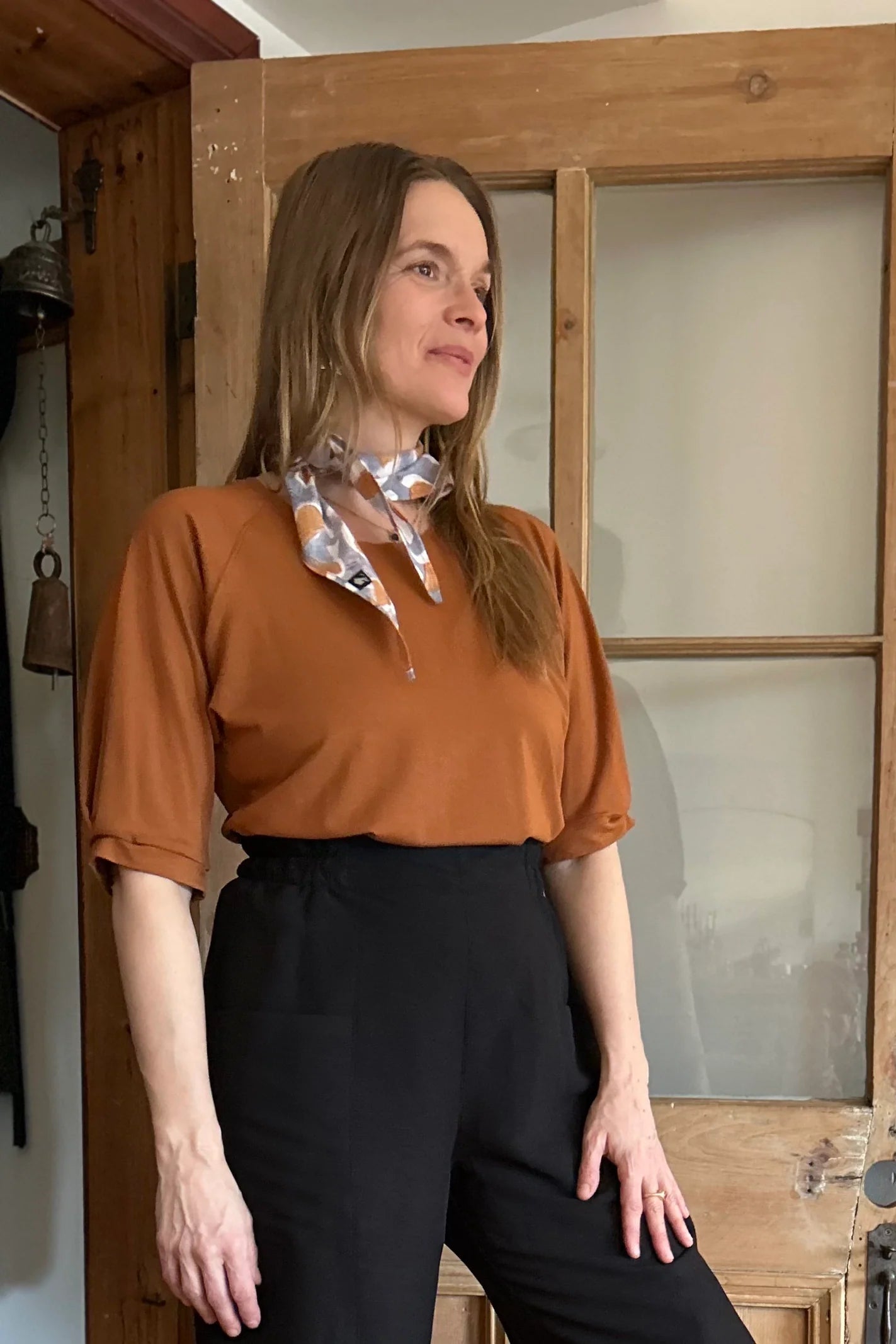 A woman wearing the Petunia Pants by Kazak in Black, wide legged cropped pants with a high waist that's smooth in front with elastic at the back, and large patch pockets. She is wearing them with an orange top and standing in front of a wooden door. 
