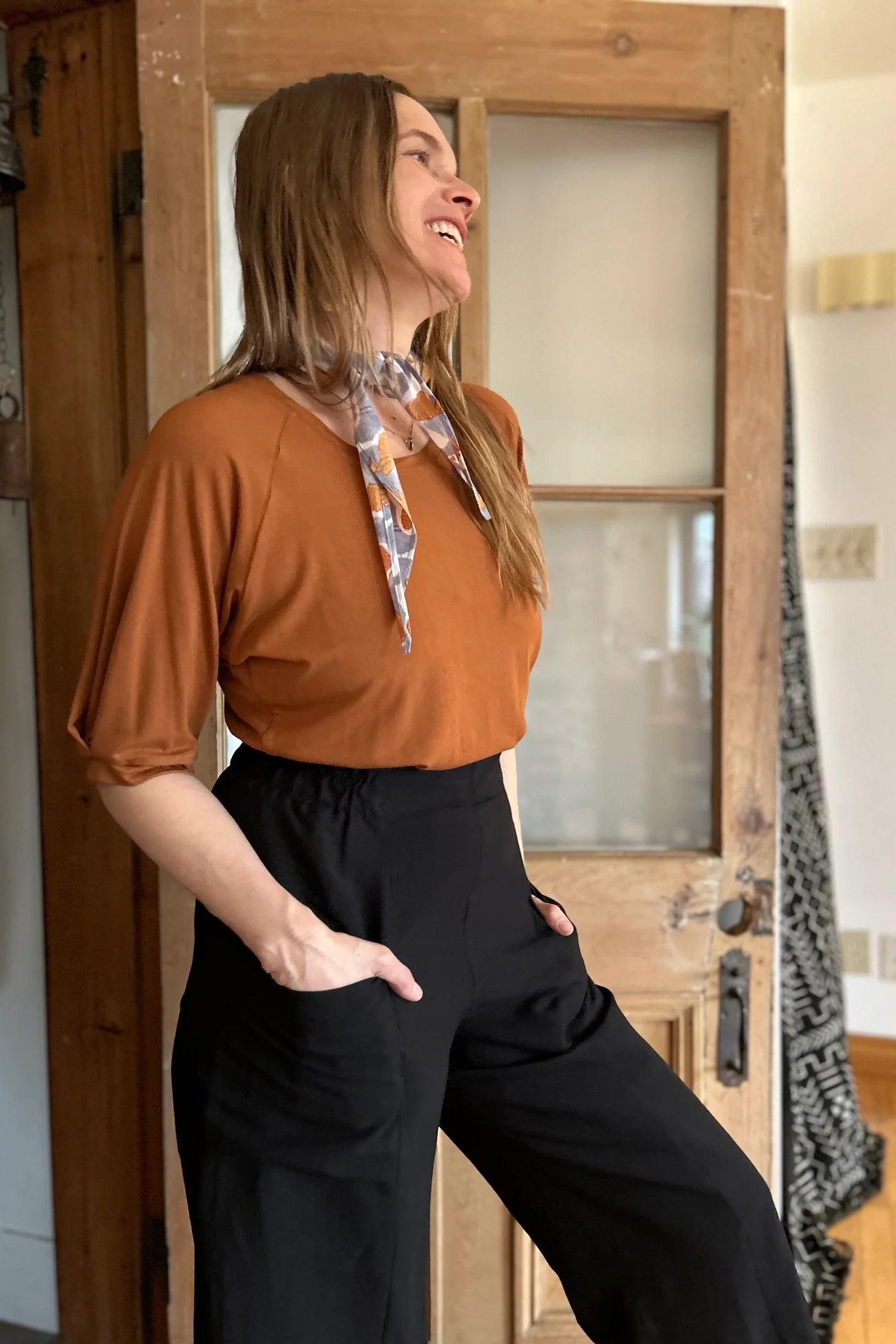 A woman wearing the Petunia Pants by Kazak in Black, wide legged cropped pants with a high waist that's smooth in front with elastic at the back, and large patch pockets. She is wearing them with an orange top and standing in front of a wooden door. 