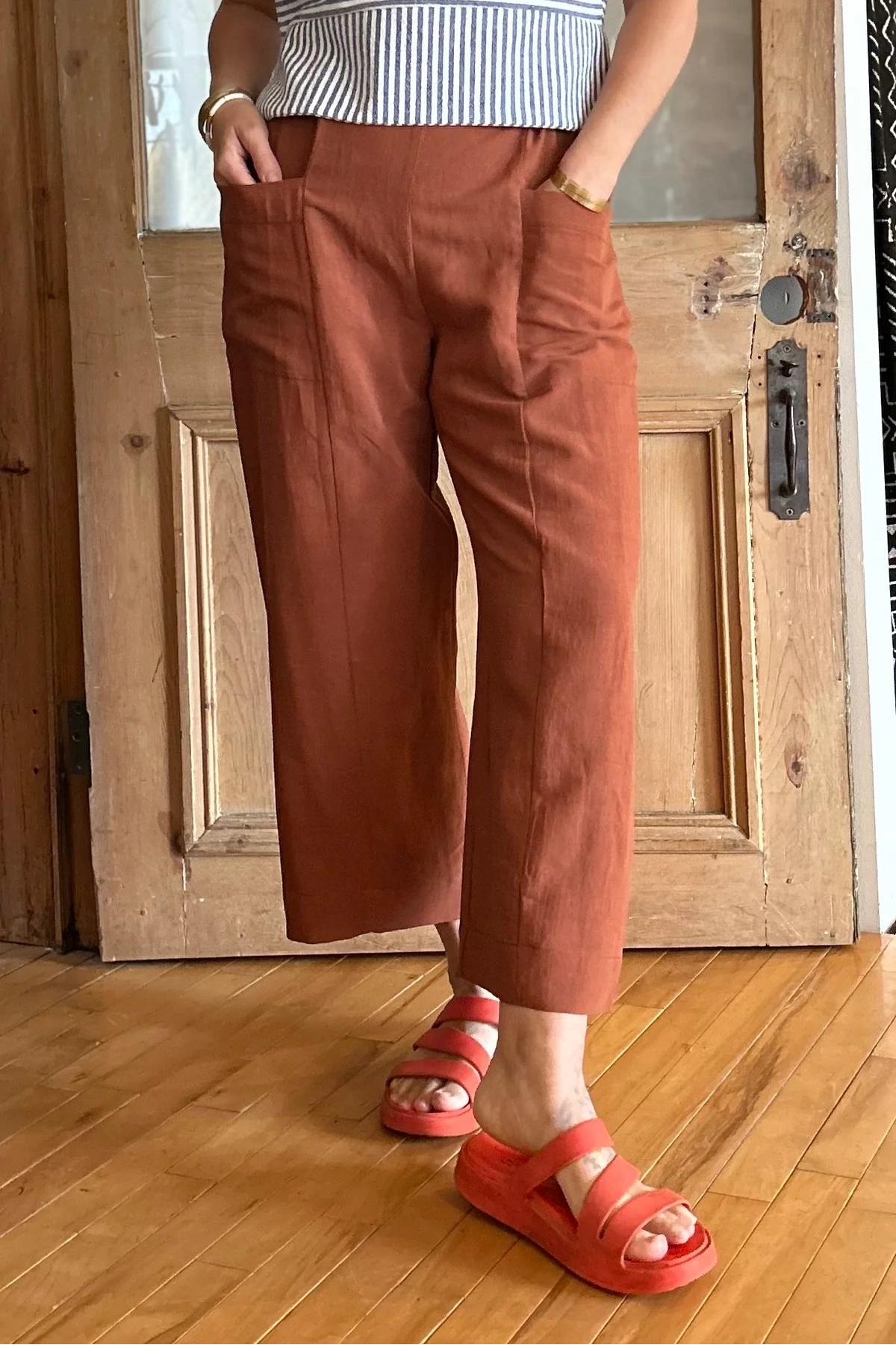 Waist down view of a woman wearing the Petunia Pants by Kazak in Terracotta, wide legged cropped pants with a high waist that's smooth in front with elastic at the back, and large patch pockets. She is wearing a striped top and standing in front of a wooden door. 