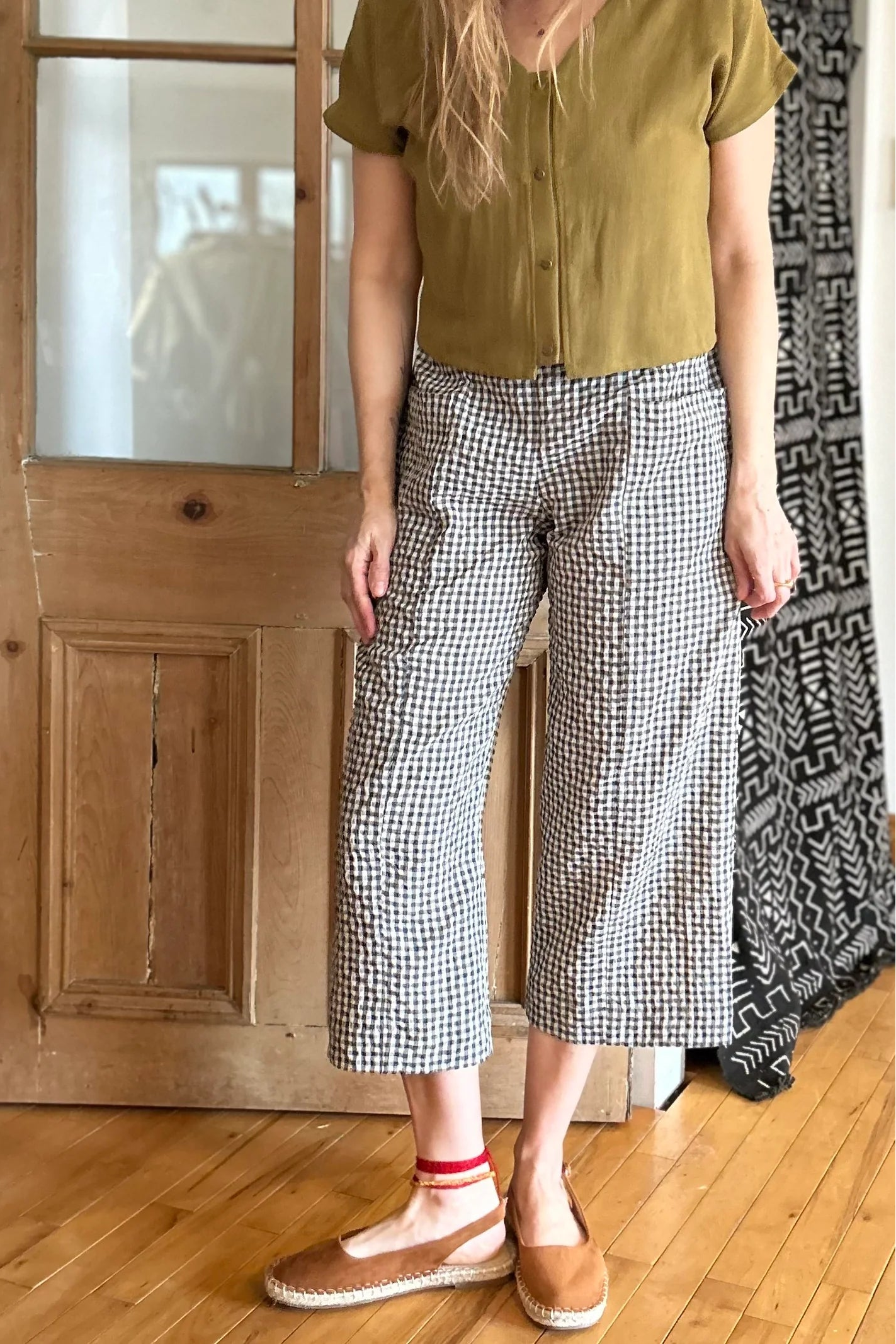Neck down view of a woman wearing the Petunia Pants by Kazak in Teal Vichy/Gingham, wide legged cropped pants with a high waist that's smooth in front with elastic at the back, and large patch pockets. She is wearing them with a green top and standing in front of a wooden door. 
