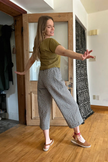 Back view of a woman wearing the Petunia Pants by Kazak in Teal Vichy/Gingham, wide legged cropped pants with a high waist that's smooth in front with elastic at the back, and large patch pockets. She is wearing them with a green top and standing in front of a wooden door. 