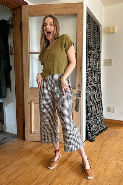 A woman wearing the Petunia Pants by Kazak in Teal Vichy/Gingham, wide legged cropped pants with a high waist that's smooth in front with elastic at the back, and large patch pockets. She is wearing them with a green top and standing in front of a wooden door. 