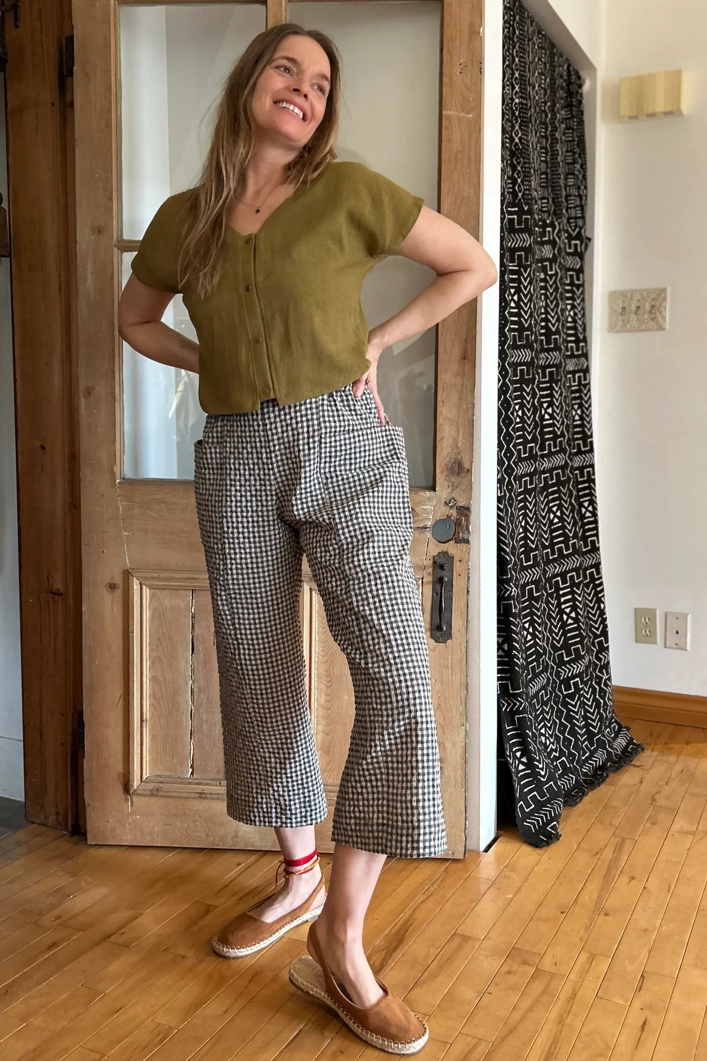 A woman wearing the Petunia Pants by Kazak in Teal Vichy/Gingham, wide legged cropped pants with a high waist that's smooth in front with elastic at the back, and large patch pockets. She is wearing them with a green top and standing in front of a wooden door. 