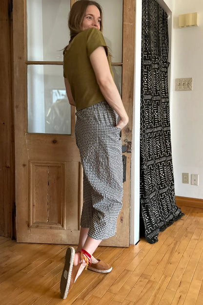 Side view of a woman wearing the Petunia Pants by Kazak in Teal Vichy/Gingham, wide legged cropped pants with a high waist that's smooth in front with elastic at the back, and large patch pockets. She is wearing them with a green top and standing in front of a wooden door. 