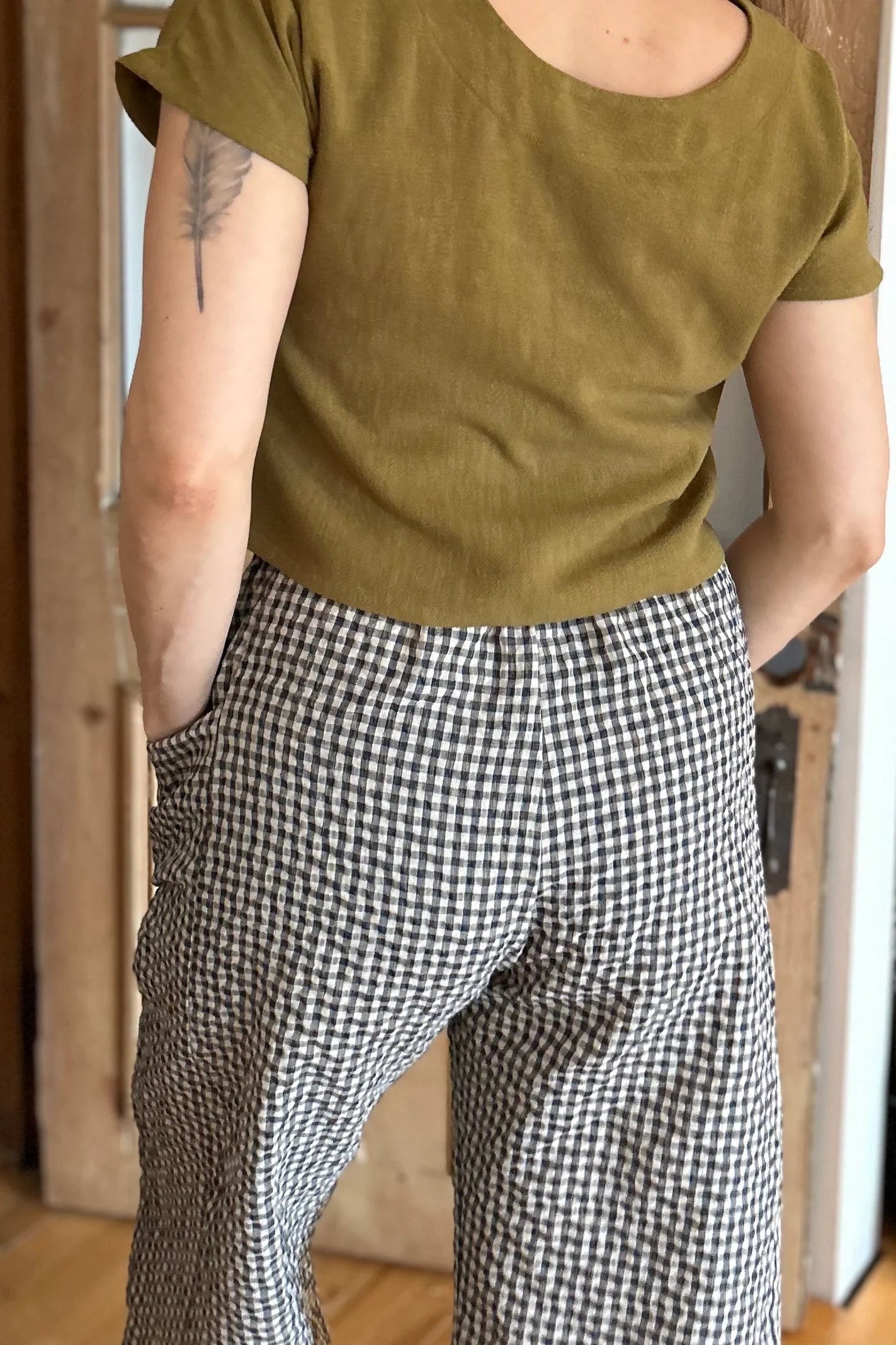 Back view of a woman wearing the Petunia Pants by Kazak in Teal Vichy/Gingham, wide legged cropped pants with a high waist that's smooth in front with elastic at the back, and large patch pockets. She is wearing them with a green top and standing in front of a wooden door. 