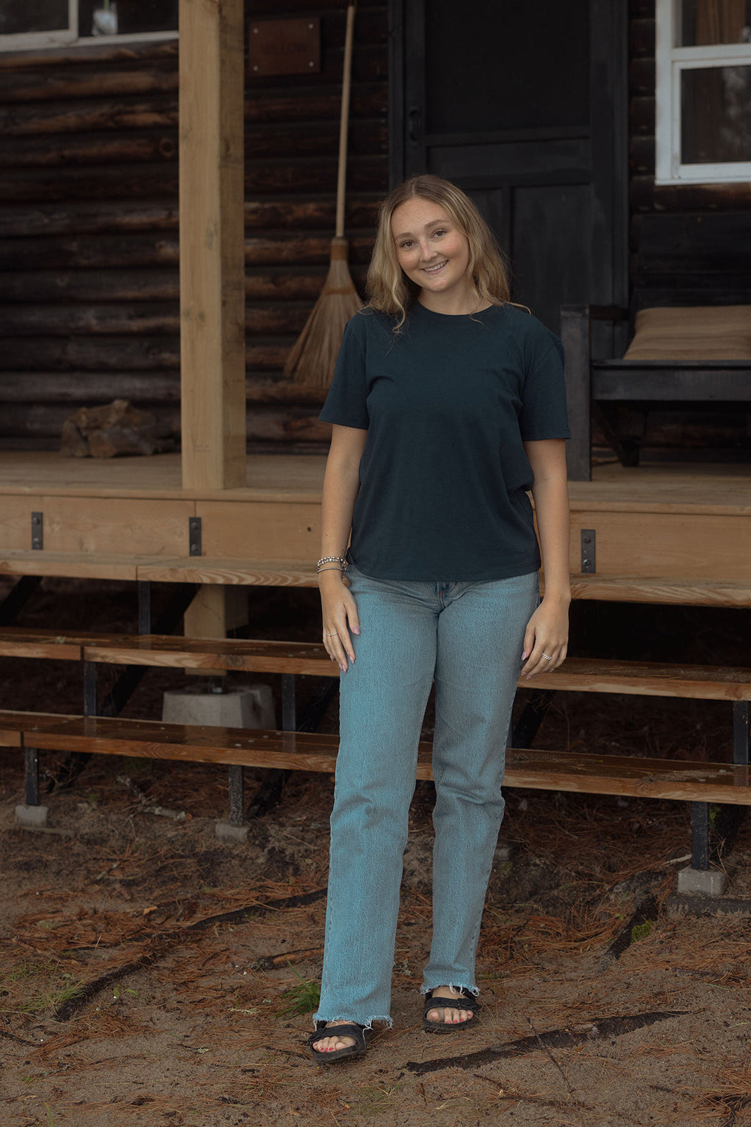 A woman wearing the Prairie Tee by Blondie in Poppy with jeans, standing on in front of a log cabin