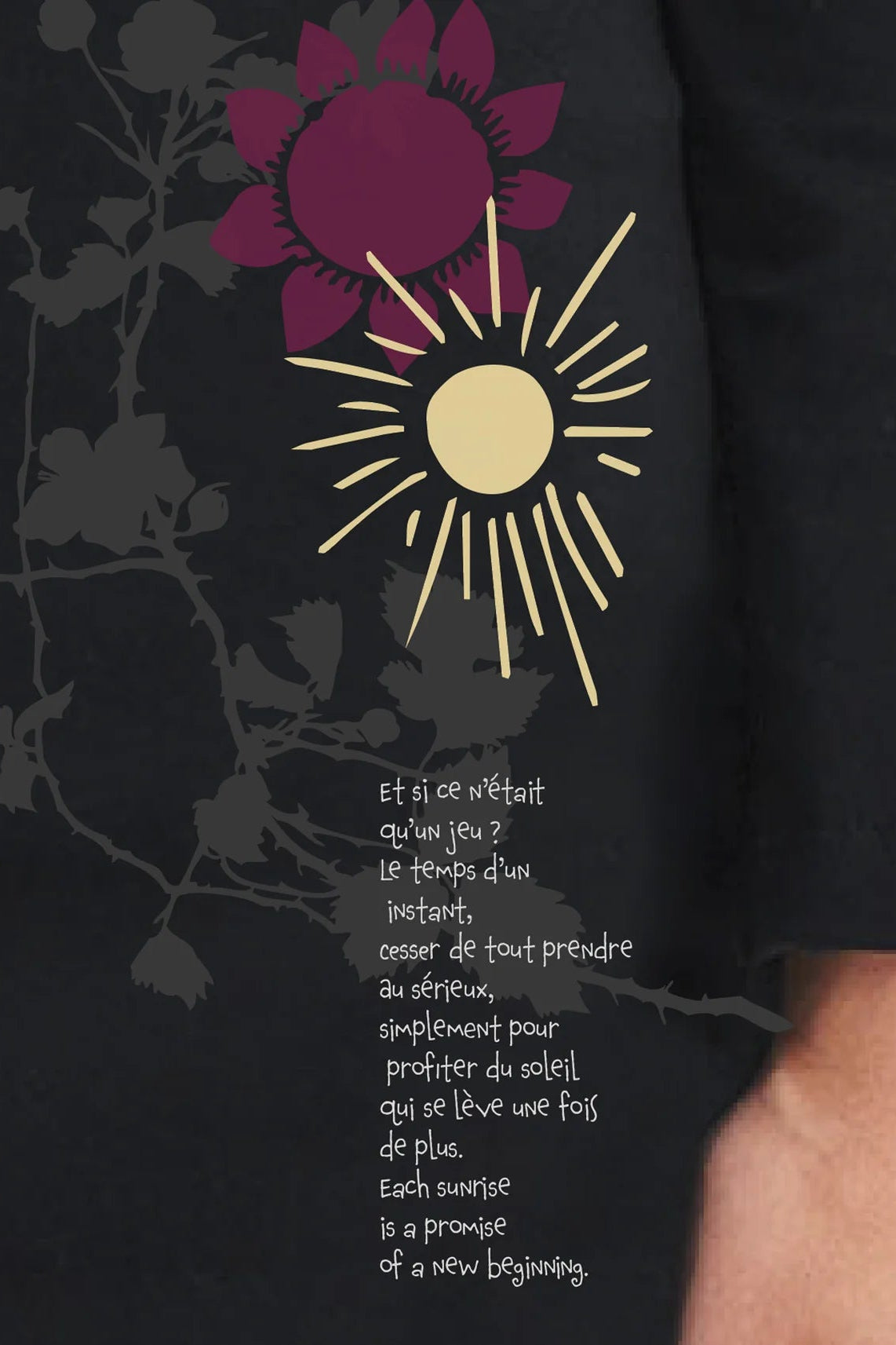 Close up of the pink and yellow nature design and French quote on the Black Oléana Tunic Dress by Message Factory. The quote reads:  “Et si ce n’était qu’un jeu ? Le temps d’un instant, cesser de tout prendre au sérieux, simplement pour profiter du soleil qui se lève une fois de plus. Each sunrise is a promise of a new beginning.” It's written in white script. 
