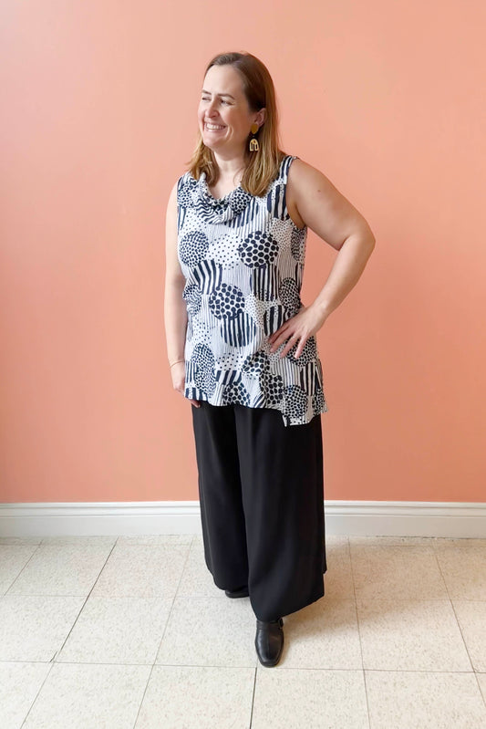 Daphne is wearing the Ebony Tunic by Pure Essence, a sleeveless tunic with a cowl neck and angled hem in a white and Indigo print. She is wearing it with black pants and standing in front of a coral wall. 