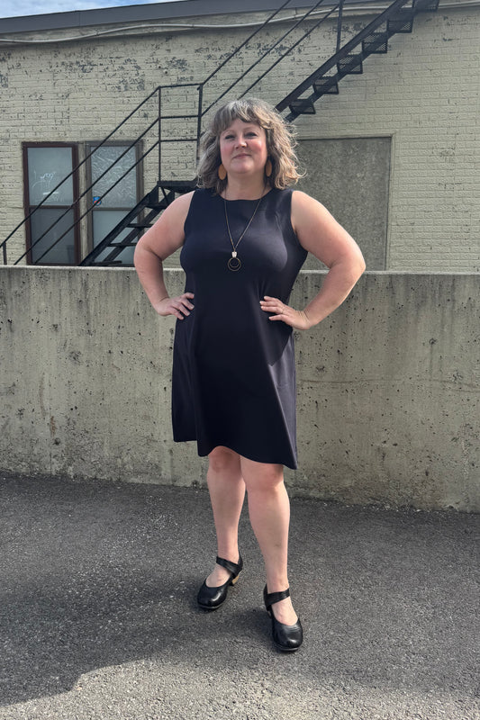 A woman wearing the Felicity Dress by Pure Essence in Navy, a sleeveless dress with a round neck, an a-line shape, and a knee length hem. She is standing in front of a brick building. 