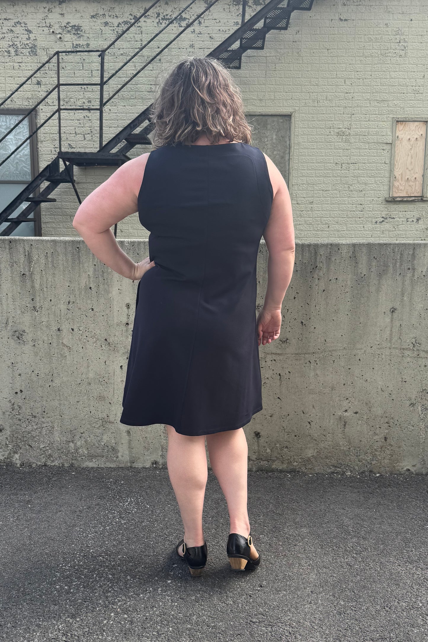 Back view of a woman wearing the Felicity Dress by Pure Essence in Navy, a sleeveless dress with a round neck, an a-line shape, and a knee length hem. She is standing in front of a brick building. 