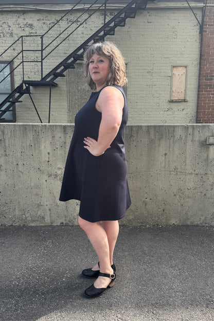 Side view of a woman wearing the Felicity Dress by Pure Essence in Navy, a sleeveless dress with a round neck, an a-line shape, and a knee length hem. She is standing in front of a brick building. 