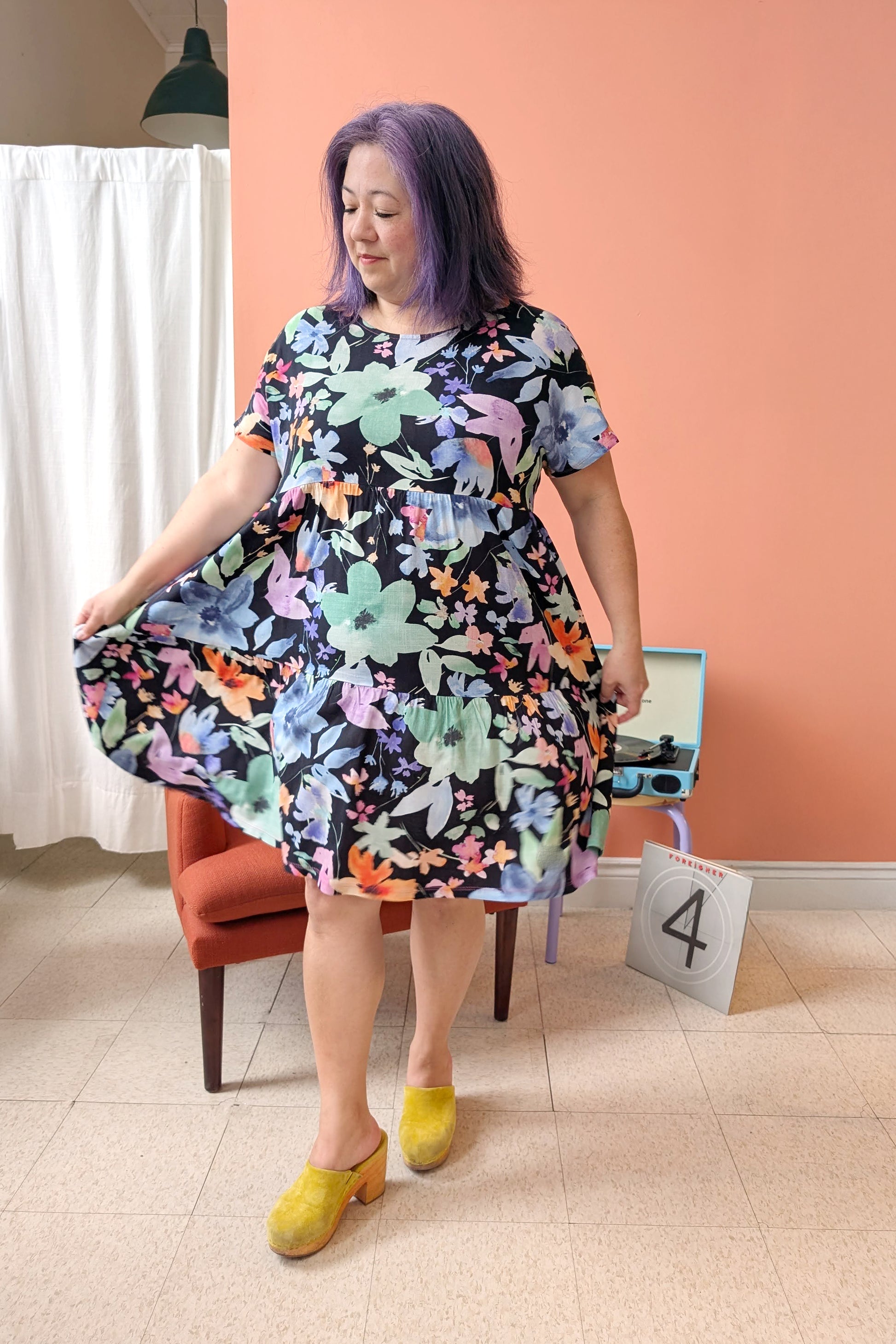 Woman wearing the Grayson Dress by Pure Essence in Moonlit Bloom, a knee-length baby doll style dress with a tiered skirt and short sleeves. She is standing in a room with a peach wall and white curtain