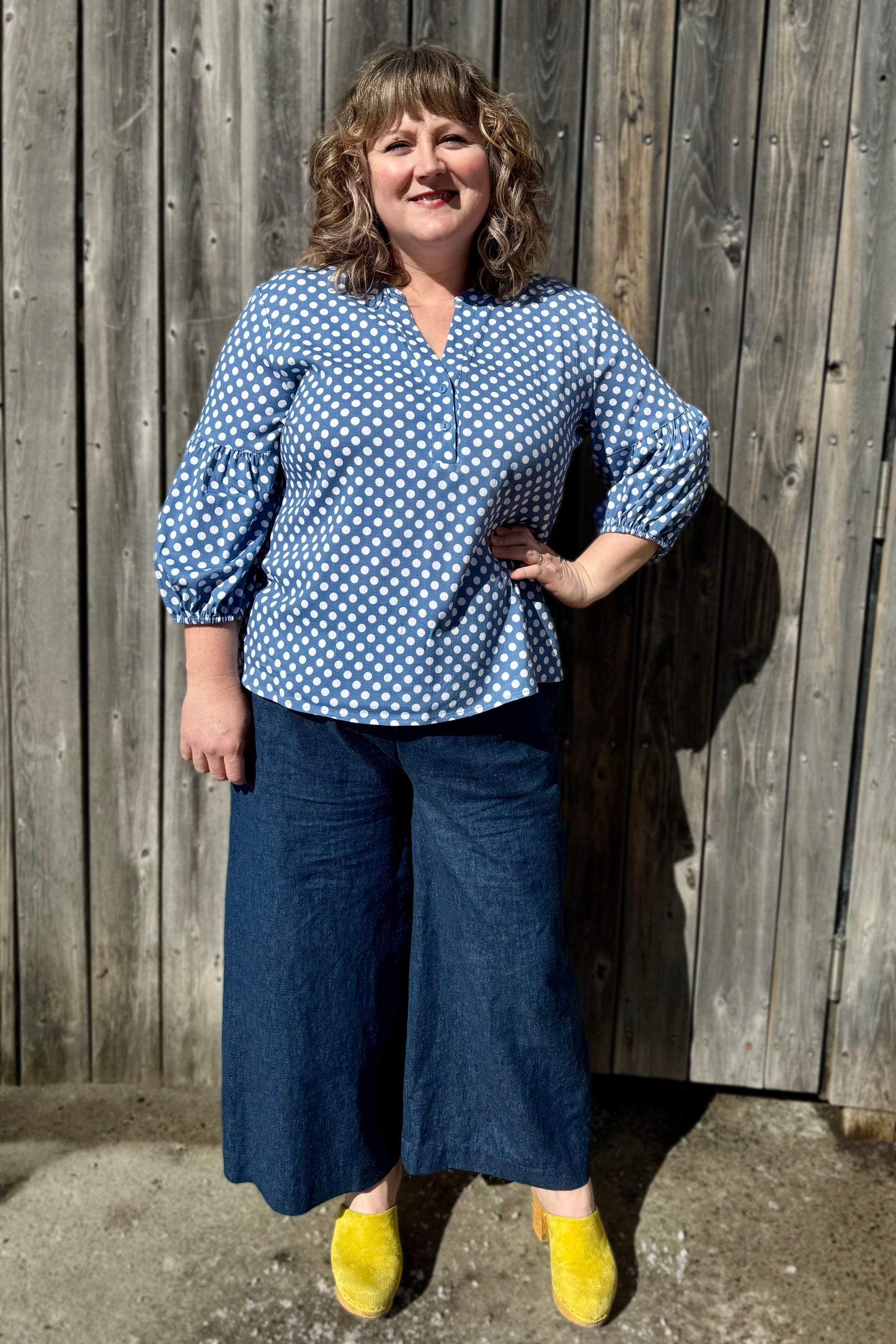 A woman wearing the Grace Blouse by Pure Essence in Chambray Dot, with a v-placket neck with buttons and 7/8 gathered sleeves. She is wearing it with denim pants and standing against a wooden fence.