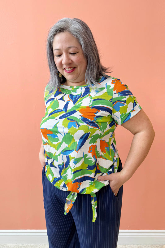 Christina is wearing the Ellen Tunic by Pure Essence in Blue/Cilantro, a short-sleeved top with a round neck and a tie detail at the waist. She is wearing it with the Eloise Pants in Ink pinstripe and is standing in front of a coral background. 