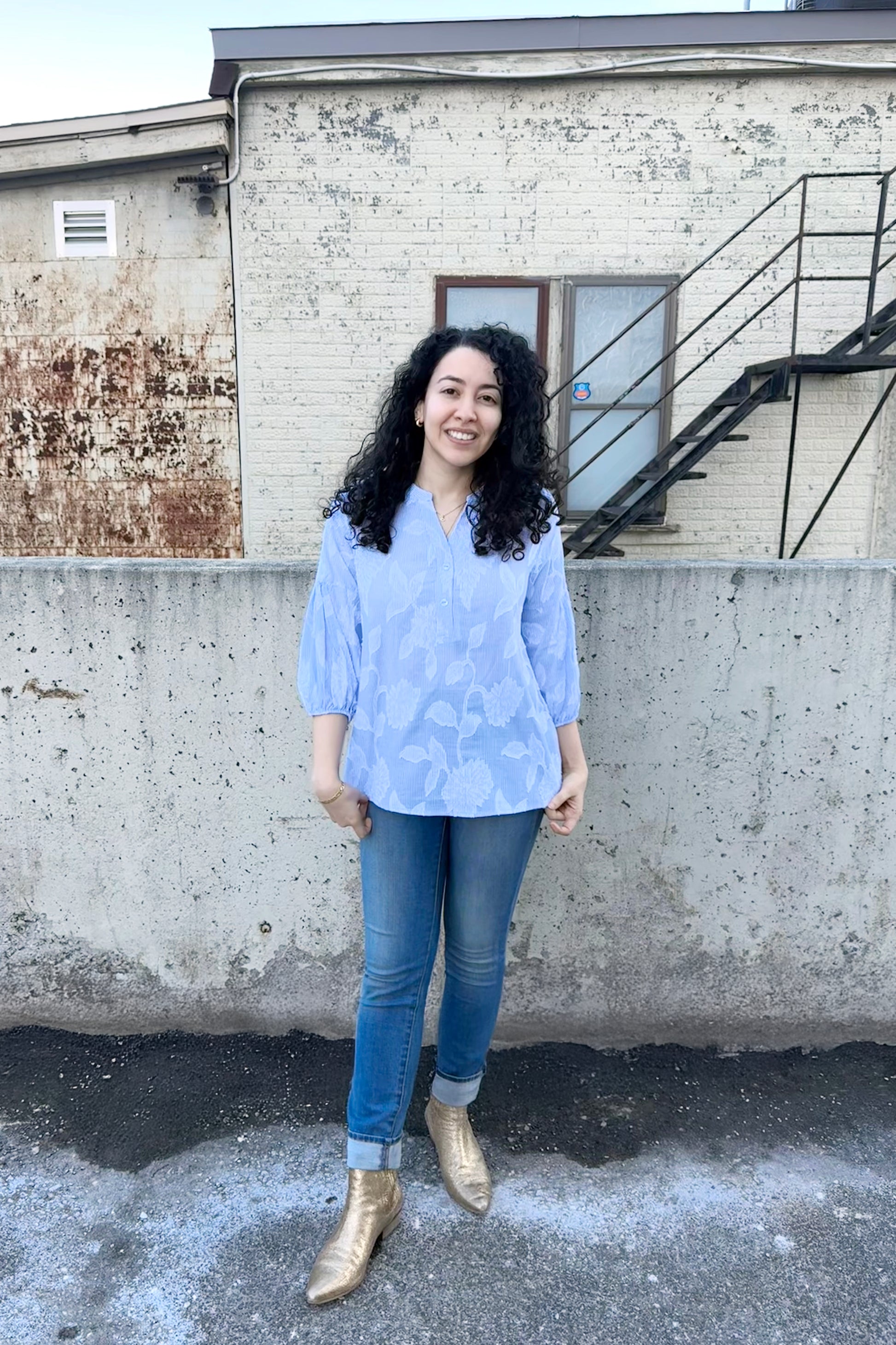 A woman wearing the Grace Blouse by Pure Essence in Blue and White, with a v-placket neck with buttons and 7/8 gathered sleeves. She is wearing it with denim pants and standing outside by a brick building.
