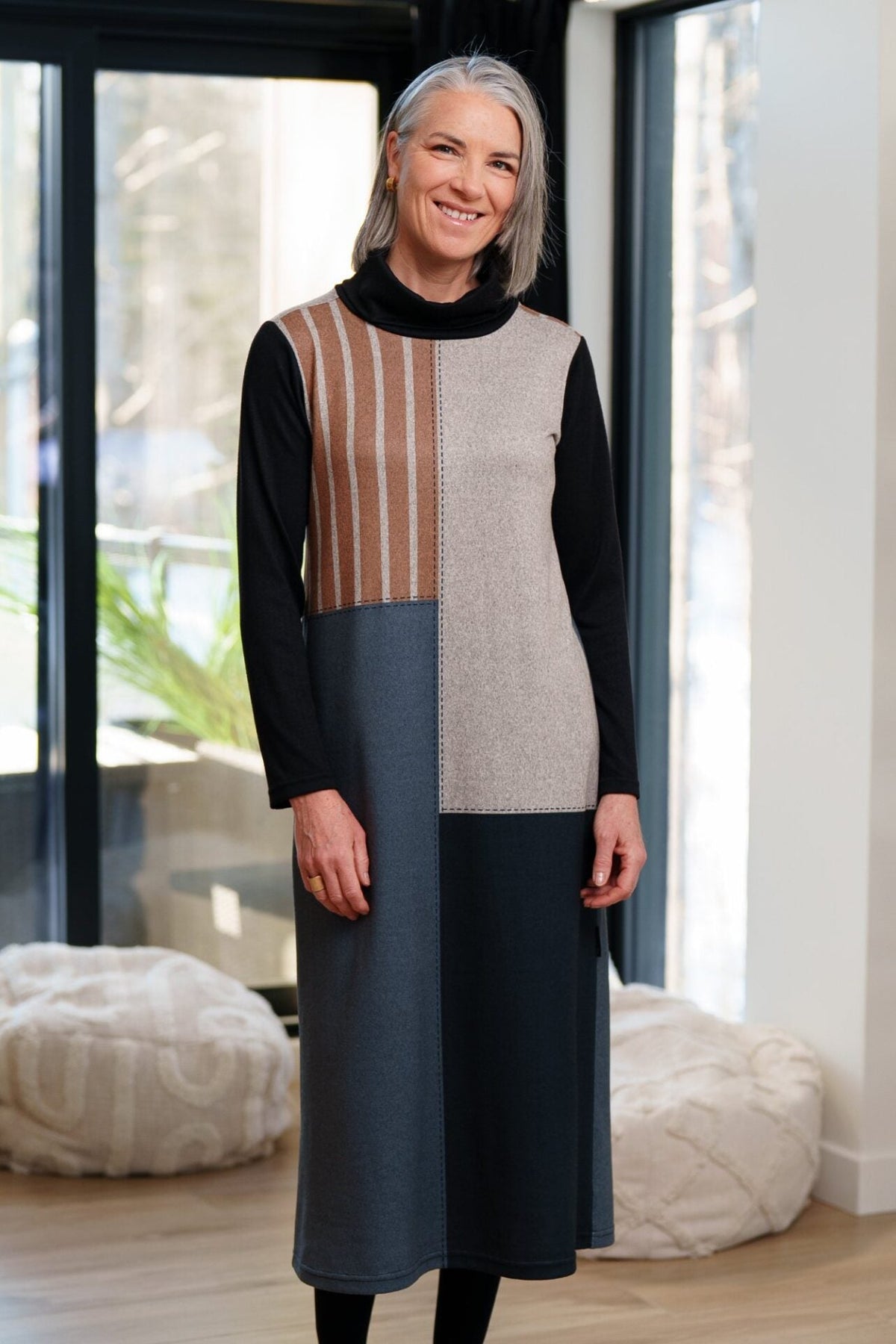 A woman wearing the Bonaventure Dress by Rien ne se Perd, a colour-blocked turtleneck sweater dress with panels of solid grey and black and brown stripes. She is standing in a room with bean bag chairs behind her. 