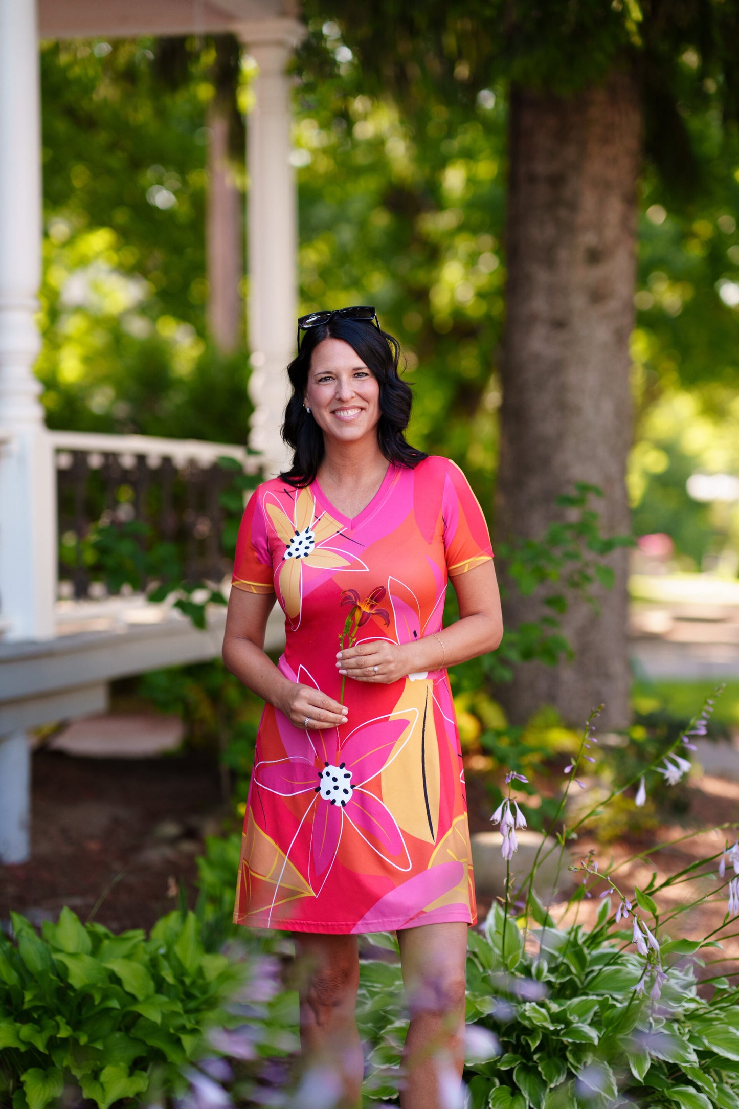 A woman wearing the Aperol Dress from Rien ne se Perd, a short flared dress with a V-neck, short sleeves, and a colourful Pink floral pattern She is standing in a garden setting.