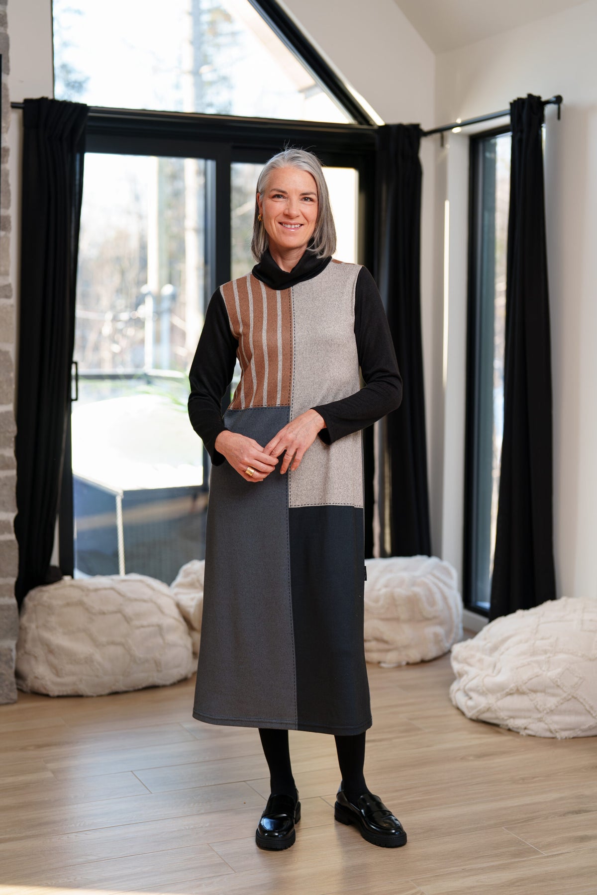 A woman wearing the Bonaventure Dress by Rien ne se Perd, a colour-blocked turtleneck sweater dress with panels of solid grey, blue and black, and brown stripes. She is standing in a room with bean bag chairs behind her. 
