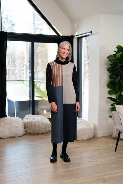 A woman wearing the Bonaventure Dress by Rien ne se Perd, a colour-blocked turtleneck sweater dress with panels of solid grey, blue and black, and brown stripes. She is standing in a room with bean bag chairs behind her. 