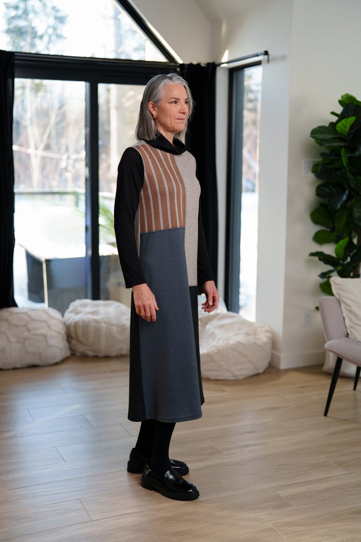 Side view of A woman wearing the Bonaventure Dress by Rien ne se Perd, a colour-blocked turtleneck sweater dress with panels of solid grey, blue and black, and brown stripes. She is standing in a room with bean bag chairs behind her. 