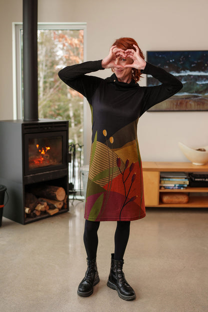 A woman wearing the Cornouiller Dress by Rien ne se Perd in Paysage print, featuring textured black fabric on the upper bodice and long sleeves and a landscape print on the body. It has a cowl neck with a slit and coconut button detail. She is standing in a room with a fireplace. 