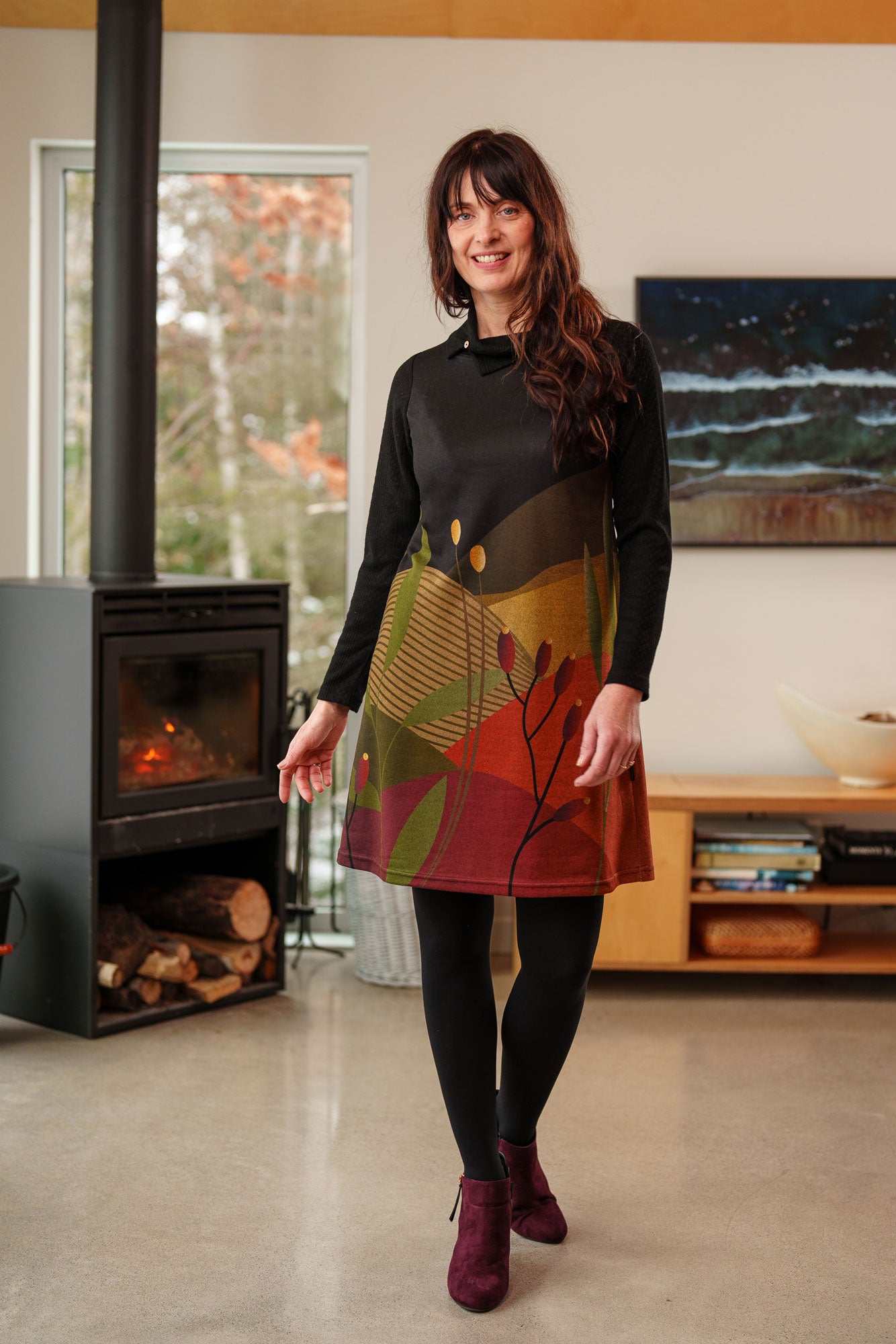 A woman wearing the Cornouiller Dress by Rien ne se Perd in Paysage print, featuring textured black fabric on the upper bodice and long sleeves and a landscape print on the body. It has a cowl neck with a slit and coconut button detail. She is standing in a room with a fireplace. 