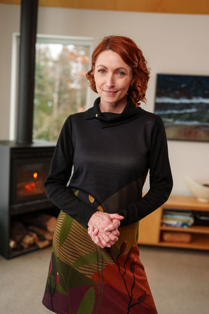 3/4 view of a woman wearing the Cornouiller Dress by Rien ne se Perd in Paysage print, featuring textured black fabric on the upper bodice and long sleeves and a landscape print on the body. It has a cowl neck with a slit and coconut button detail. She is standing in a room with a fireplace. 