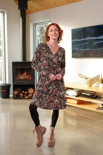 A woman wearing the Epilobe Dress by Rien ne se Perd in Fall Paisley, a classic wrap dress with a tie at the waist, delicate pleats at the shoulders, and a hem that hits above the knee. She is standing in a living room with a fireplace and TV.