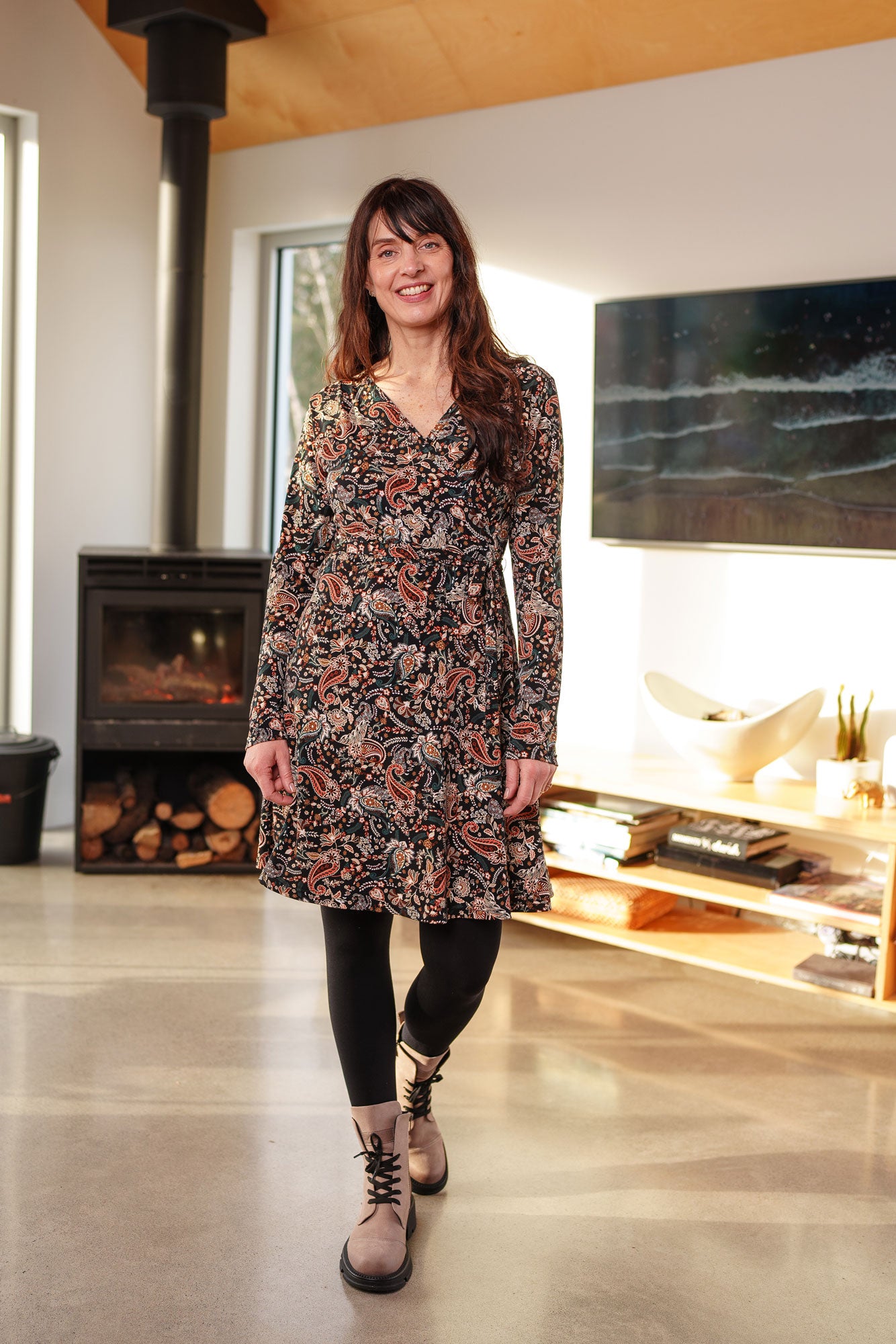 A woman wearing the Epilobe Dress by Rien ne se Perd in Fall Paisley, a classic wrap dress with a tie at the waist, delicate pleats at the shoulders, and a hem that hits above the knee. She is standing in a living room with a fireplace and TV.