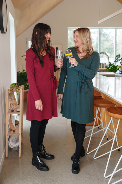 Two women wearing the Labrador Dress by Rien ne se Perd, one in Emerald and one in Garnet, a semi-fitted dress with a v-neck, long sleeves, tie detail at the side waist, and above the knee length. They are standing in a kitchen, holding glasses of wine. 