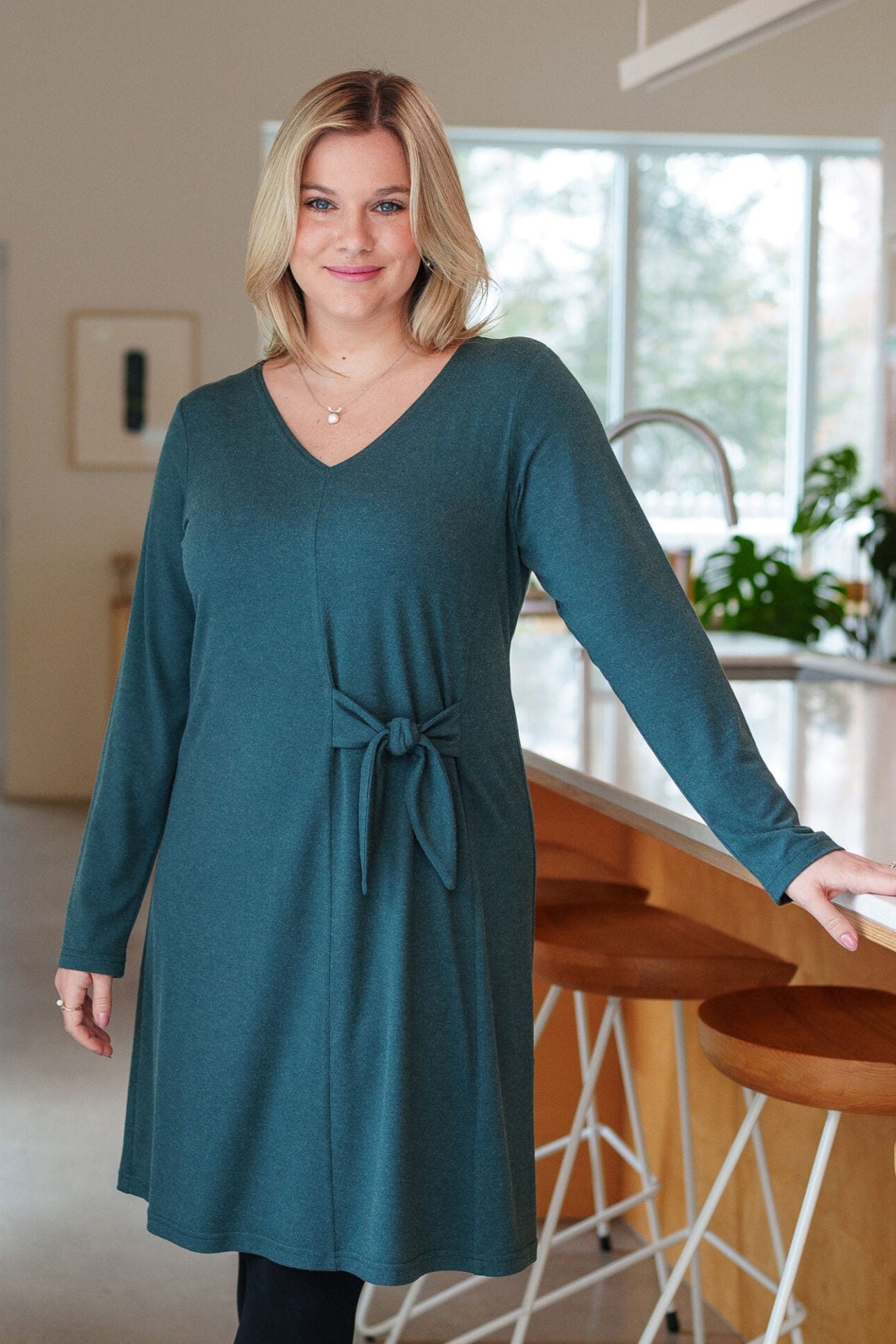 A woman wearing the Labrador Dress by Rien ne se Perd in Emerald, a semi-fitted dress with a v-neck, long sleeves, tie detail at the side waist, and above the knee length. She is standing in a kitchen. 