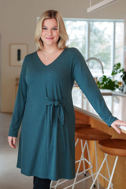 A woman wearing the Labrador Dress by Rien ne se Perd in Emerald, a semi-fitted dress with a v-neck, long sleeves, tie detail at the side waist, and above the knee length. She is standing in a kitchen. 