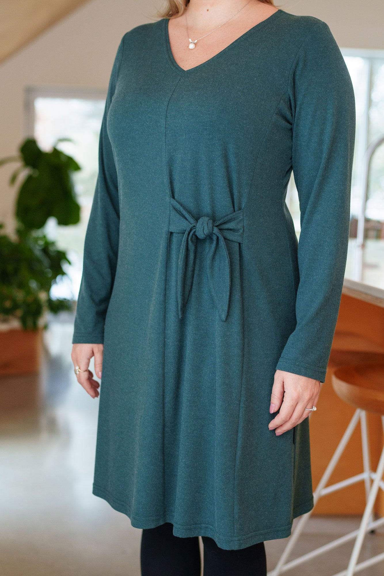 Torso view of a woman wearing the Labrador Dress by Rien ne se Perd in Emerald, a semi-fitted dress with a v-neck, long sleeves, tie detail at the side waist, and above the knee length. She is standing in a kitchen. 