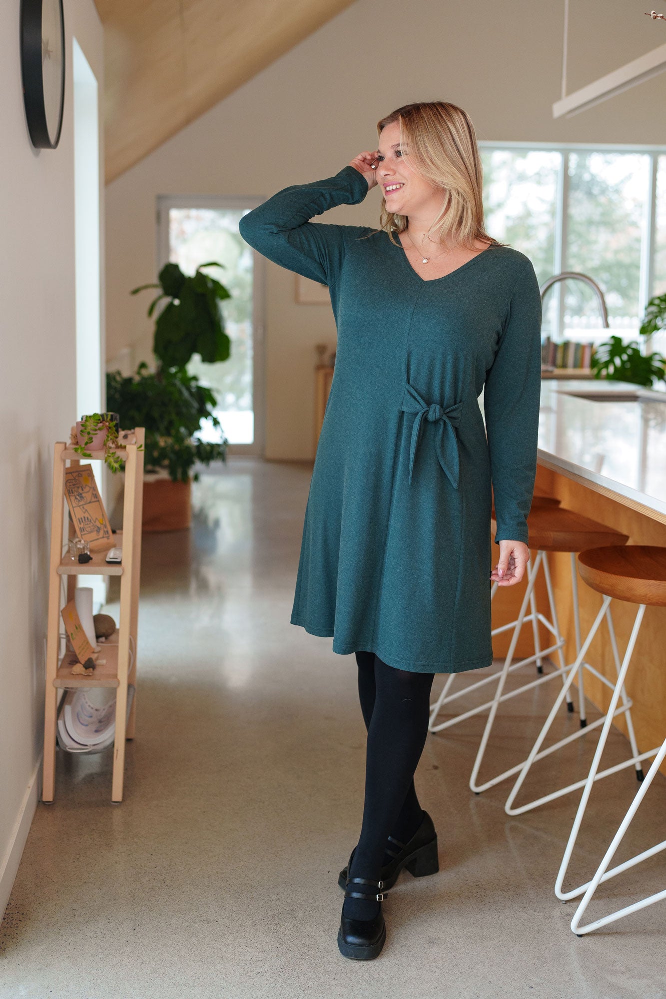 A woman wearing the Labrador Dress by Rien ne se Perd in Emerald, a semi-fitted dress with a v-neck, long sleeves, tie detail at the side waist, and above the knee length. She is standing in a kitchen. 