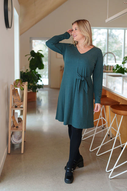 A woman wearing the Labrador Dress by Rien ne se Perd in Emerald, a semi-fitted dress with a v-neck, long sleeves, tie detail at the side waist, and above the knee length. She is standing in a kitchen. 