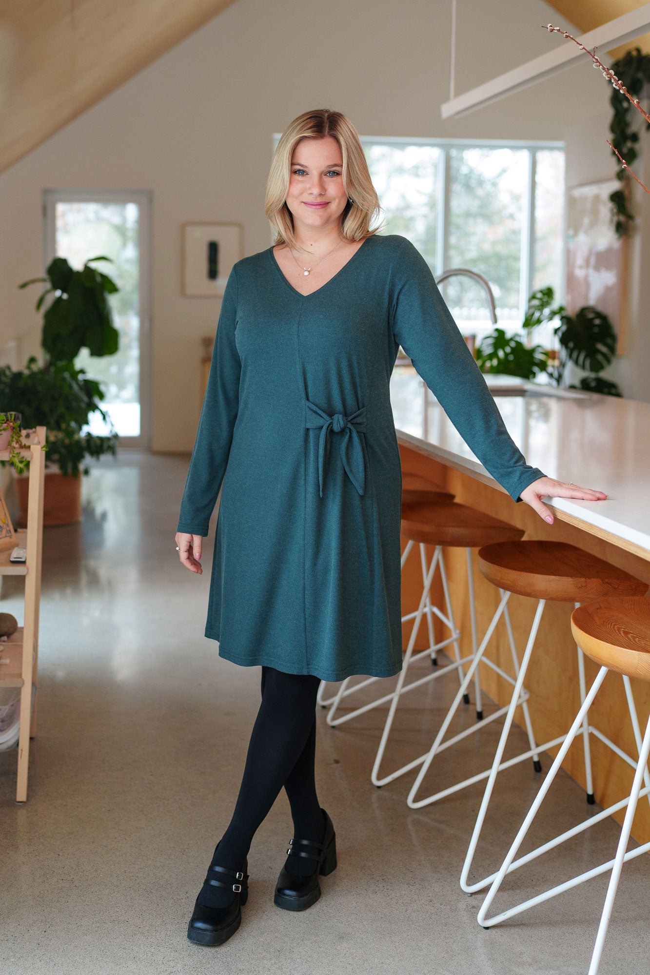 A woman wearing the Labrador Dress by Rien ne se Perd in Emerald, a semi-fitted dress with a v-neck, long sleeves, tie detail at the side waist, and above the knee length. She is standing in a kitchen. 