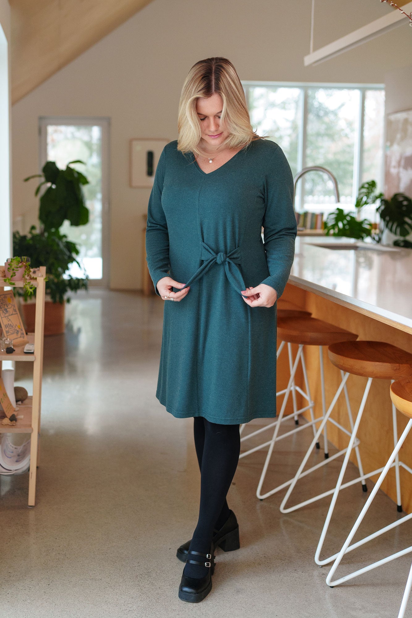 A woman wearing the Labrador Dress by Rien ne se Perd in Emerald, a semi-fitted dress with a v-neck, long sleeves, tie detail at the side waist, and above the knee length. She is standing in a kitchen. 