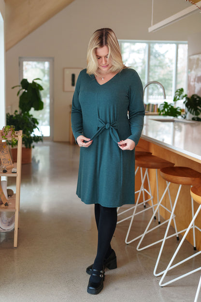 A woman wearing the Labrador Dress by Rien ne se Perd in Emerald, a semi-fitted dress with a v-neck, long sleeves, tie detail at the side waist, and above the knee length. She is standing in a kitchen. 