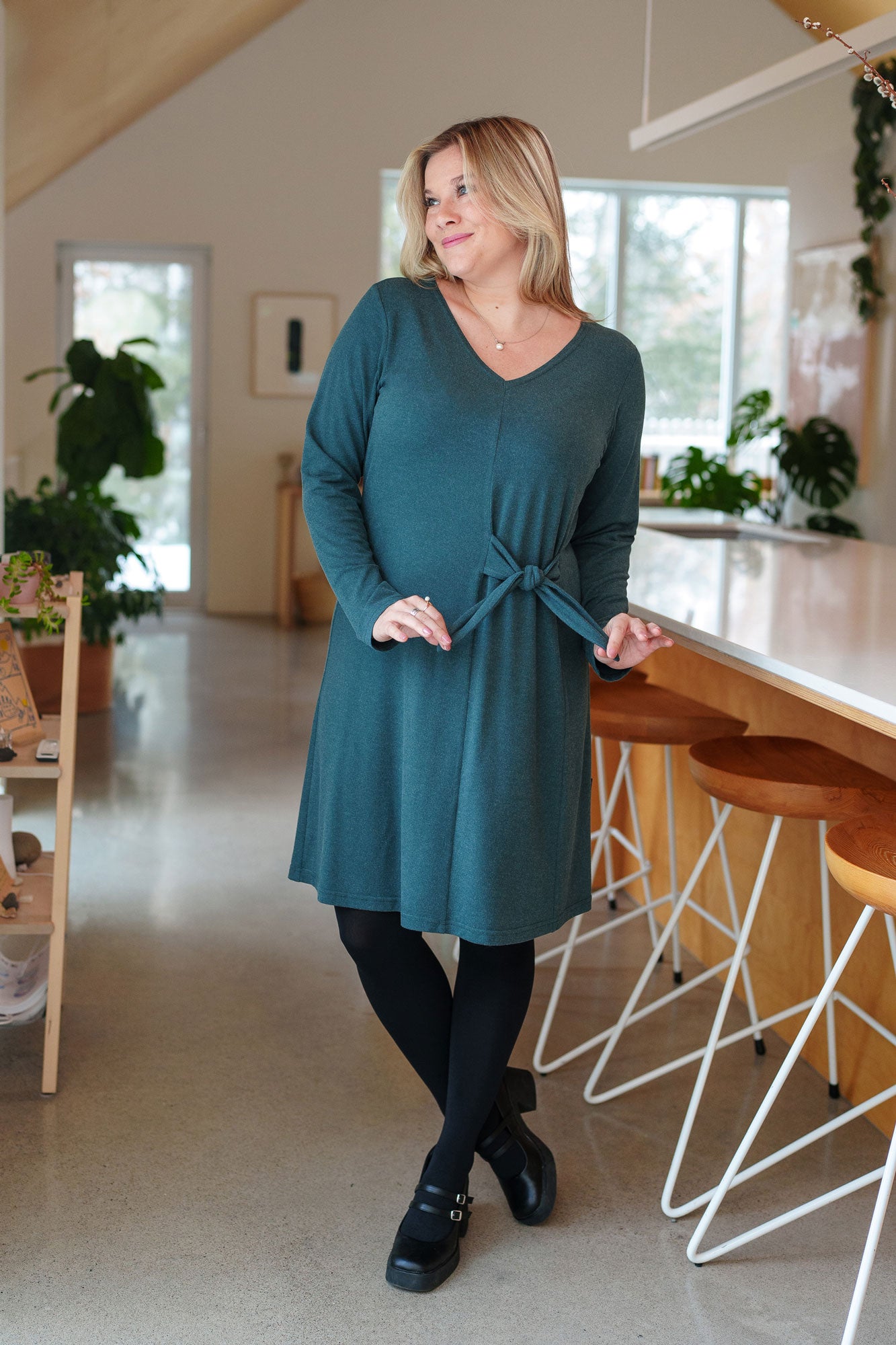 A woman wearing the Labrador Dress by Rien ne se Perd in Emerald, a semi-fitted dress with a v-neck, long sleeves, tie detail at the side waist, and above the knee length. She is standing in a kitchen. 
