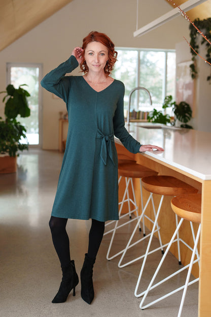 A woman wearing the Labrador Dress by Rien ne se Perd in Emerald, a semi-fitted dress with a v-neck, long sleeves, tie detail at the side waist, and above the knee length. She is standing in a kitchen. 
