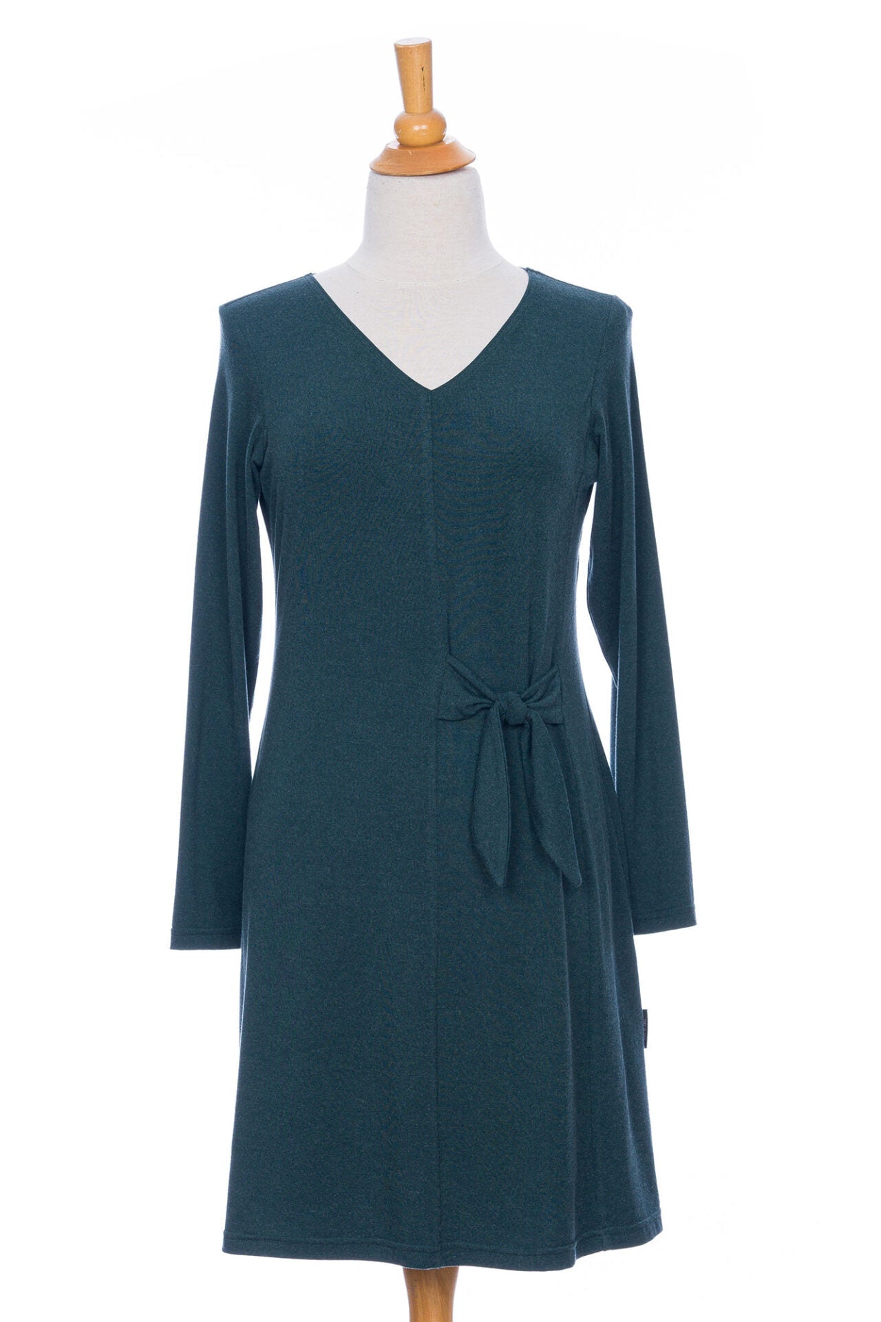 The Labrador Dress by Rien ne se Perd in Emerald, a semi-fitted dress with a v-neck, long sleeves, tie detail at the side waist, and above the knee length, is shown on a mannequin in front of a white background. 