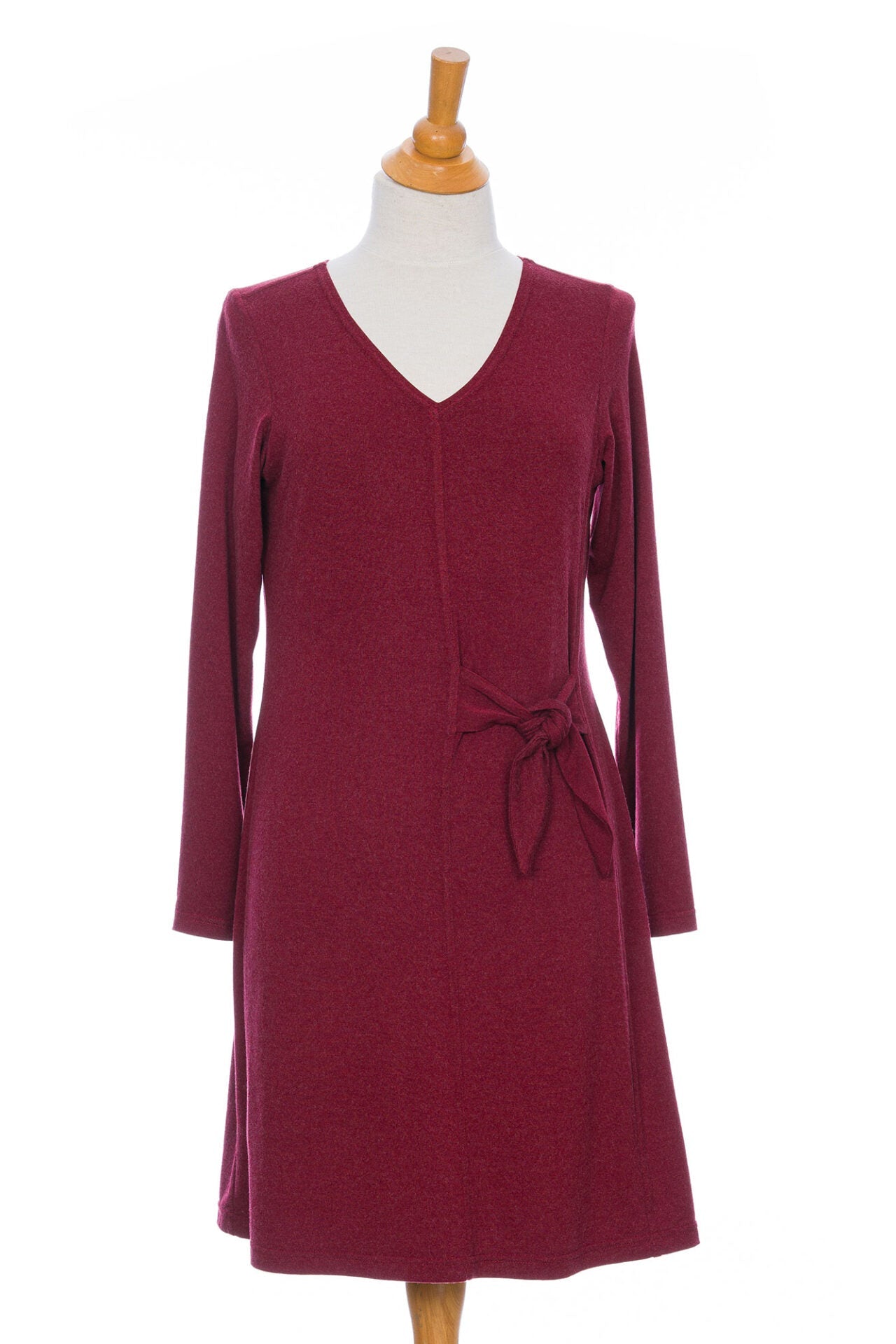 The Labrador Dress by Rien ne se Perd in Garnet, a semi-fitted dress with a v-neck, long sleeves, tie detail at the side waist, and above the knee length, is shown on a mannequin in front of a white background. 