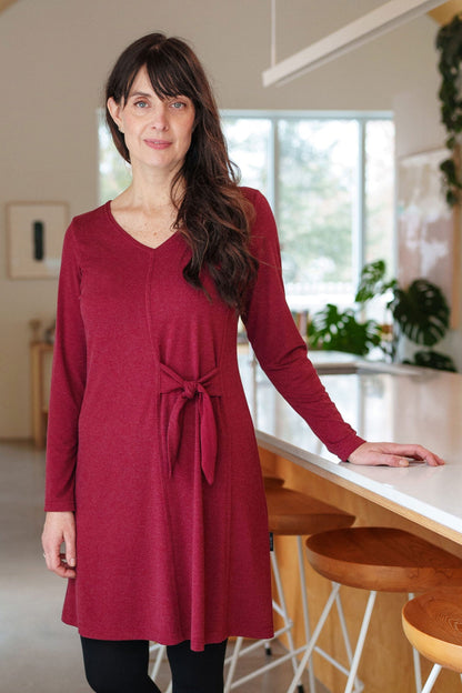A woman wearing the Labrador Dress by Rien ne se Perd in Garnet, a semi-fitted dress with a v-neck, long sleeves, tie detail at the side waist, and above the knee length. She is standing in a kitchen. 