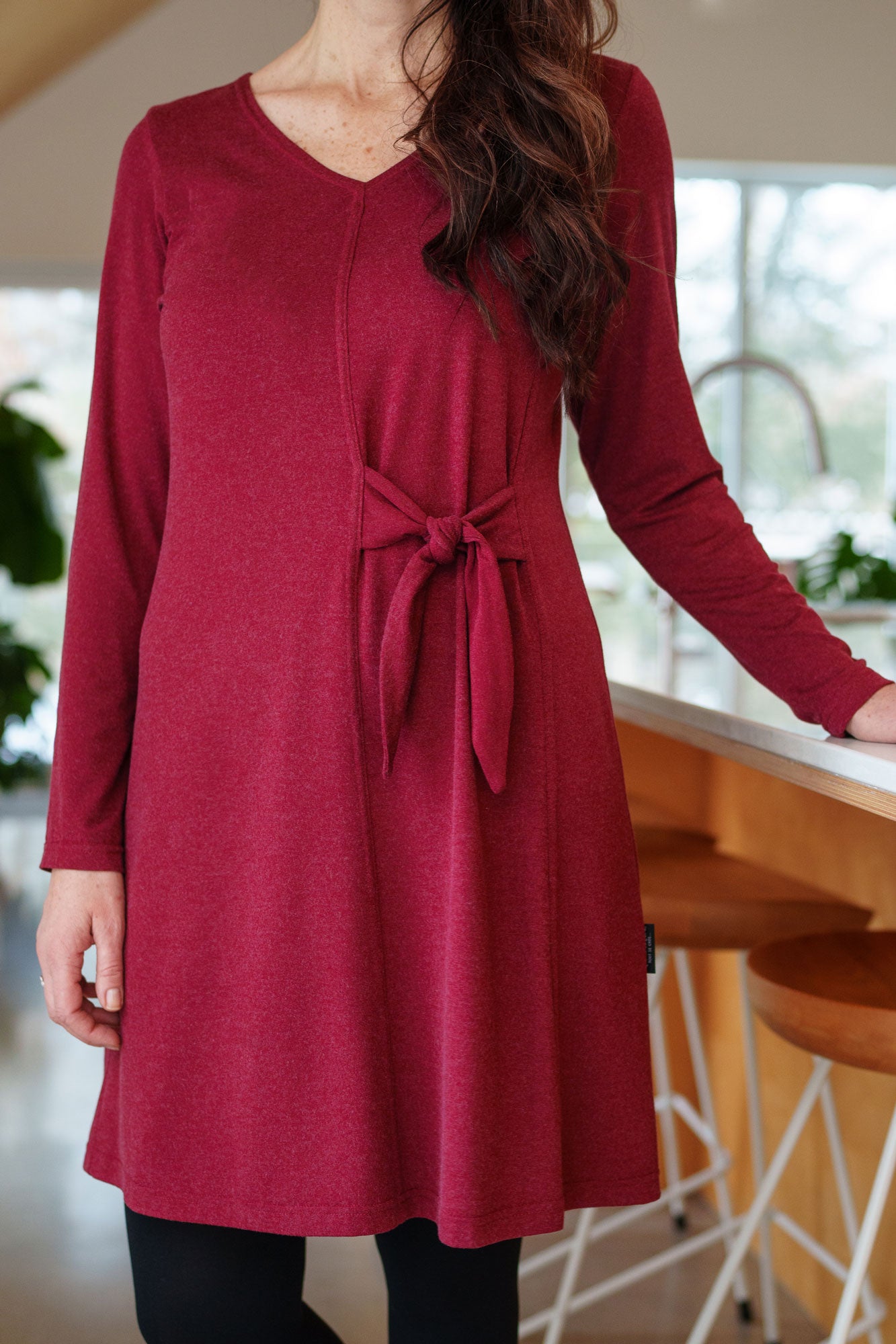 Torso view of a woman wearing the Labrador Dress by Rien ne se Perd in Garnet, a semi-fitted dress with a v-neck, long sleeves, tie detail at the side waist, and above the knee length. She is standing in a kitchen. 