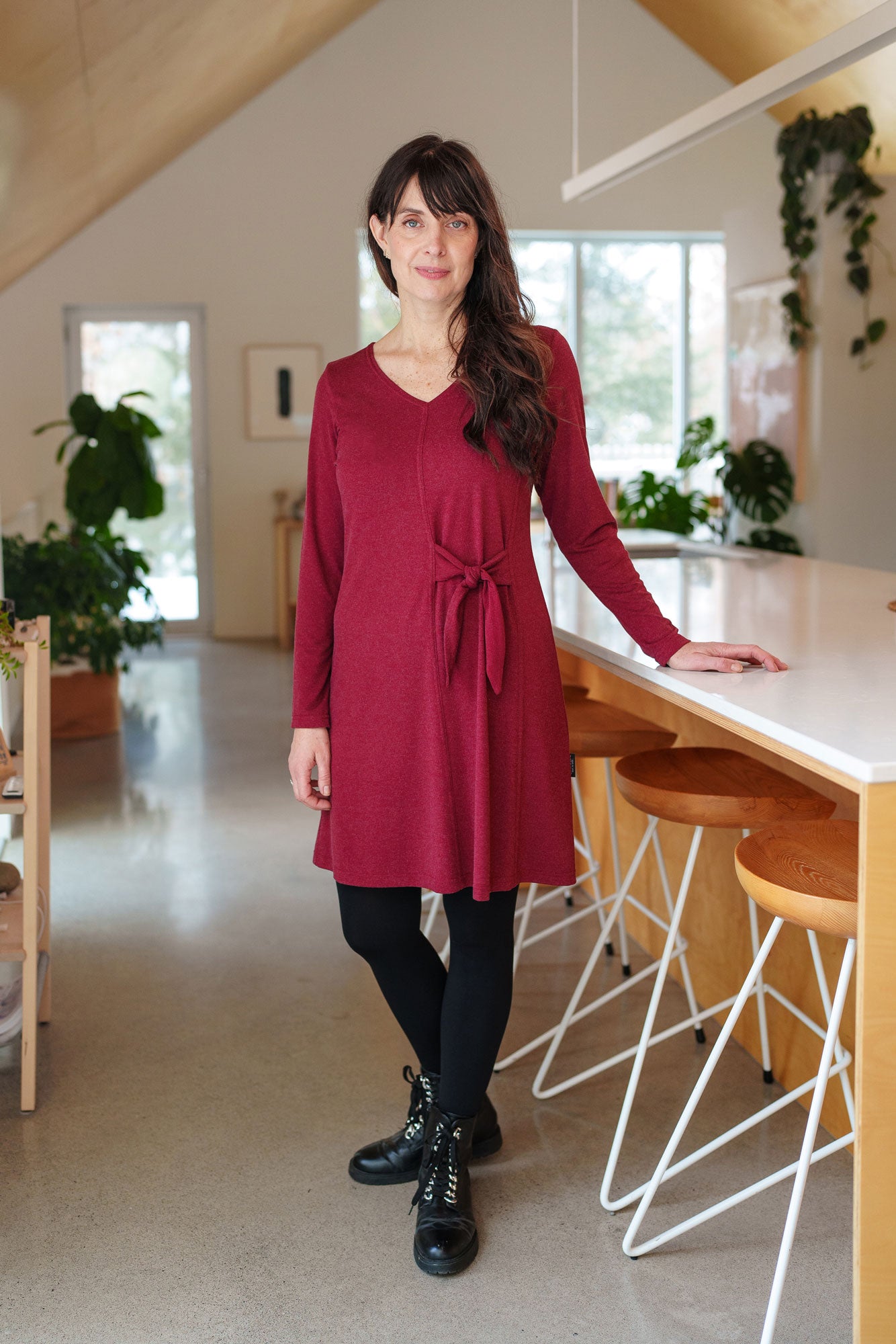 A woman wearing the Labrador Dress by Rien ne se Perd in Garnet, a semi-fitted dress with a v-neck, long sleeves, tie detail at the side waist, and above the knee length. She is standing in a kitchen. 